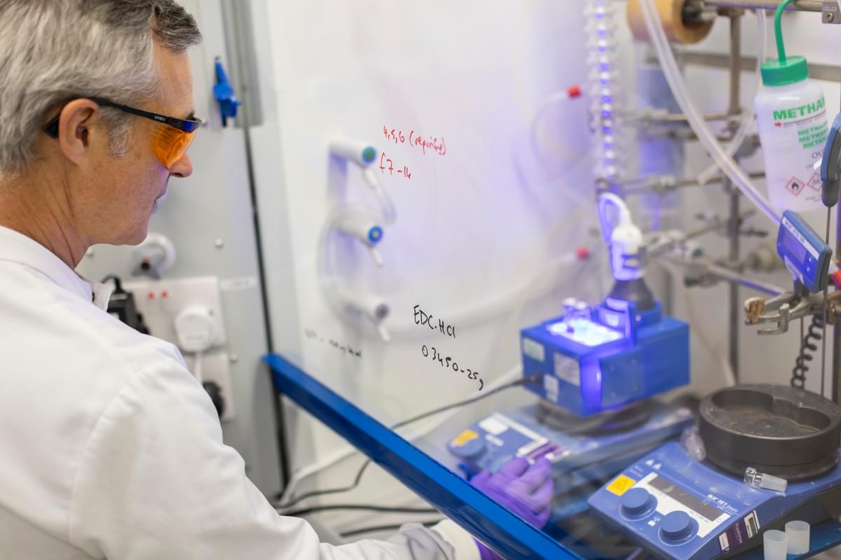 A LifeArc scientist operates chemistry equipment behind a protective screen.