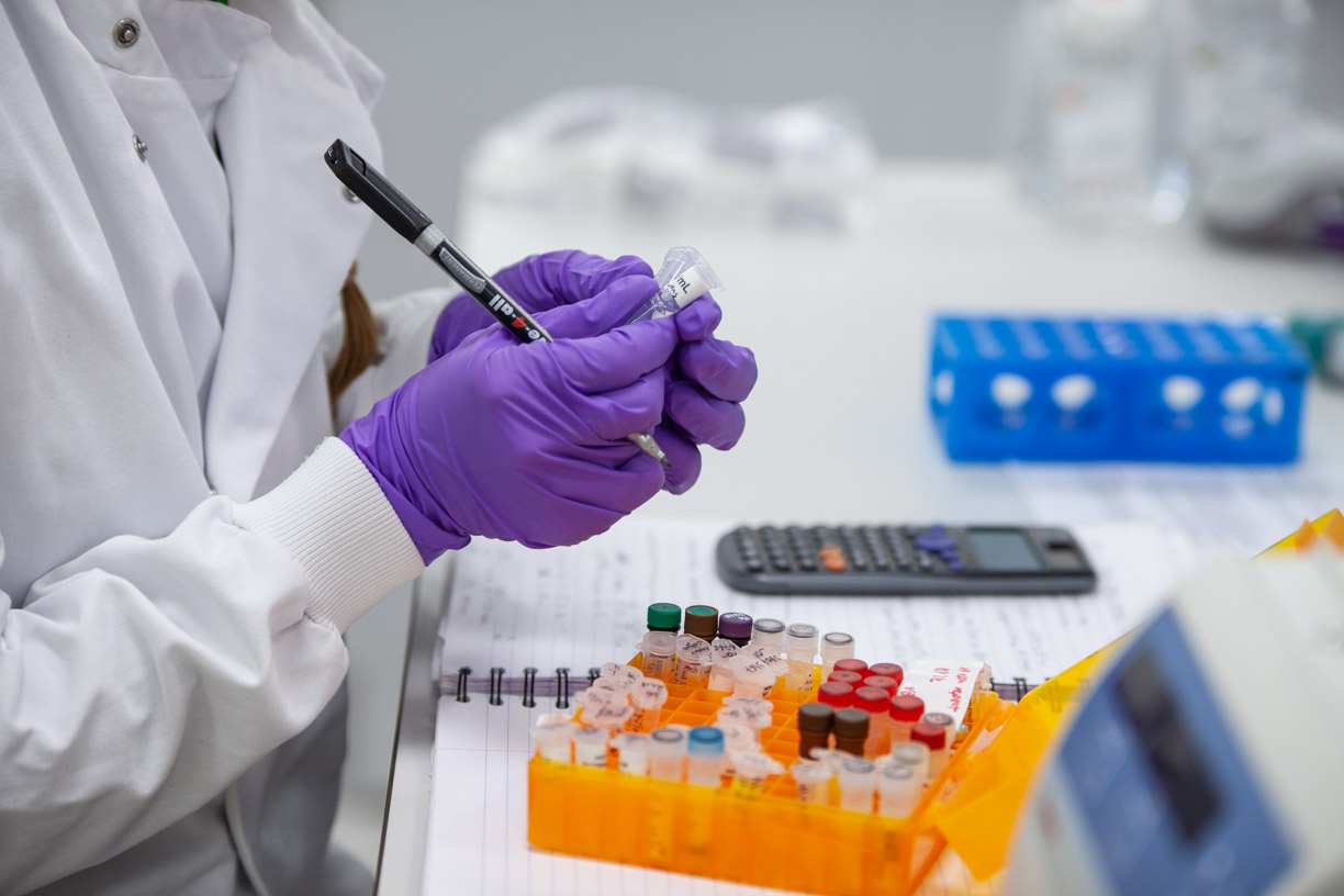 The gloved hands of a lab-coated scientist hold two vials and a pen above lab bench and a tray of vials.