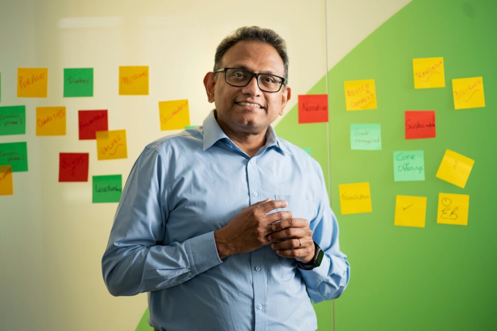 LifeArc employee, Madhu Madhusudhan, smiling standing infront of a wall with post-it notes on